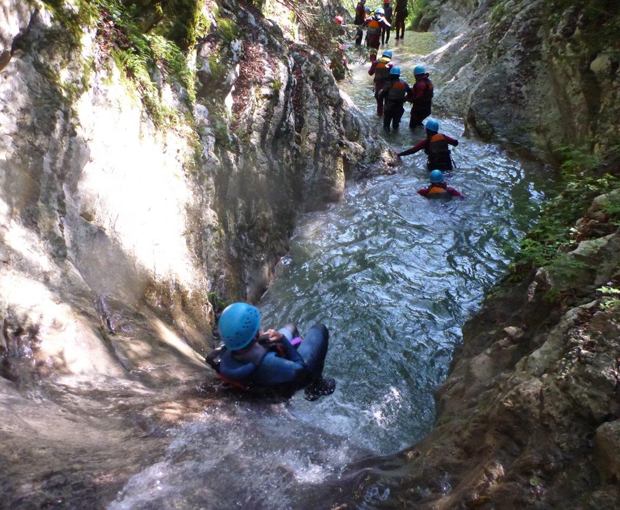 A person wearing a blue helmet slides down a narrow, rocky water channel. Others in similar gear, some standing, form a line behind, surrounded by lush greenery and sunlit cliffs.