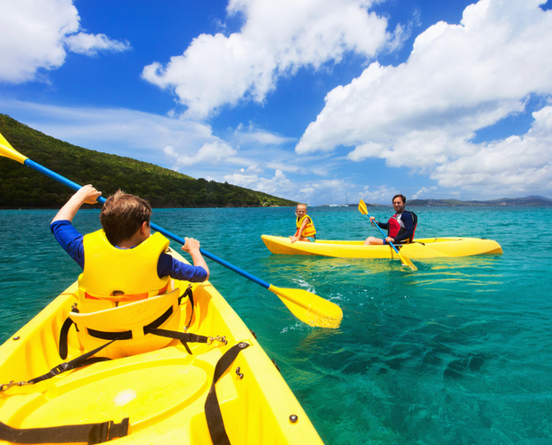 Three people in yellow kayaks paddle on clear blue water, wearing life jackets under a bright sky with fluffy clouds, surrounded by lush green hills.