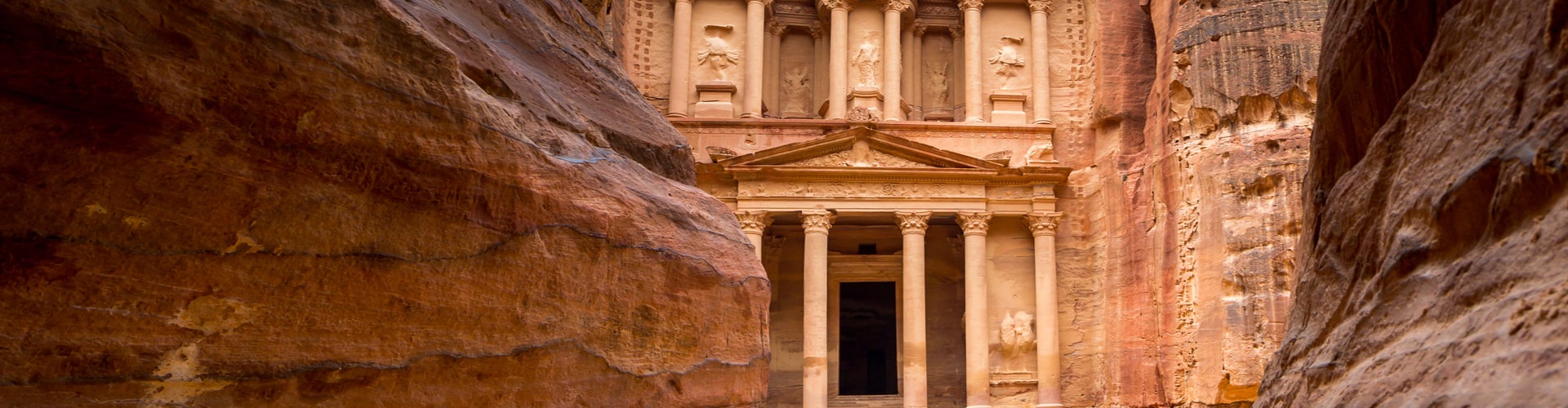 Ancient stone building with columns carved into reddish rock, viewed through a narrow canyon. The structure is intricately detailed and set within a rugged, natural landscape.
