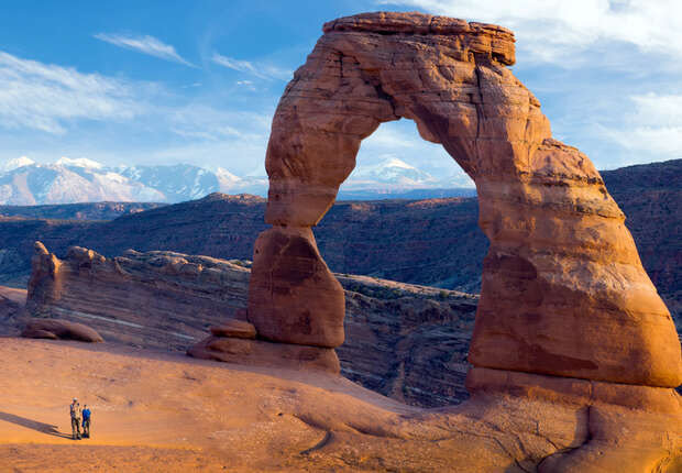 A large, naturally-formed sandstone arch stands in a desert landscape, with two people standing nearby, surrounded by distant mountain peaks and a bright, blue sky.