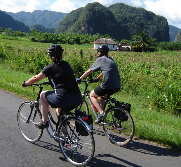 Two people ride bicycles along a rural road, surrounded by lush greenery and large hills in the background, under a clear sky.