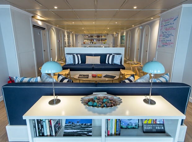 A cozy lounge features two blue sofas facing each other, book-filled shelves, and a distant bar counter. Soft lighting and blue lamps complement the striped cushions, creating a relaxed atmosphere.