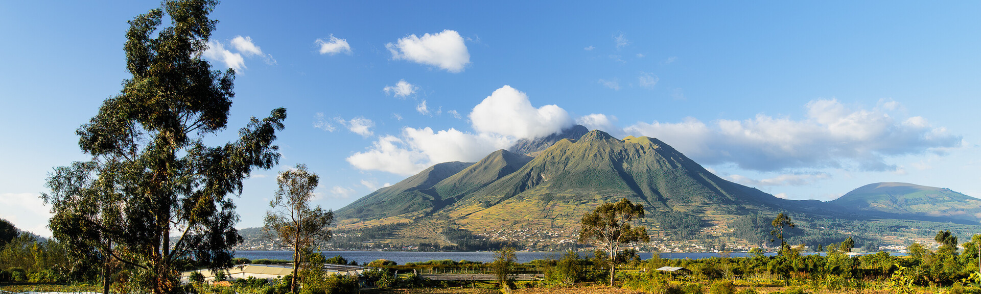 A mountain stands with its peak partially covered by clouds, surrounded by a lake, trees, and a few scattered houses under a clear blue sky.