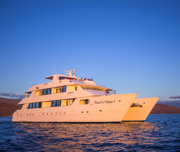 A large white yacht named "Treasure of Galapagos" floats calmly on blue ocean waters during sunset, with several people visible on deck enjoying the warm, golden light.