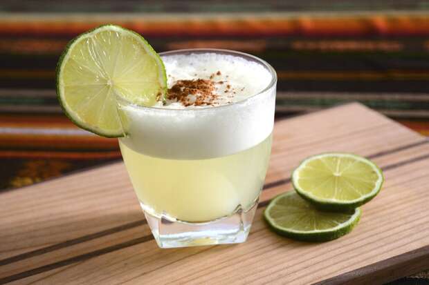 A cocktail glass holds a yellowish drink with a frothy top and spice garnish, adorned with a lime slice. It rests on a wooden board beside two lime slices, with a striped background.
