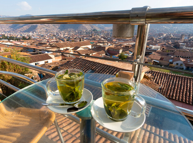 Two transparent cups filled with herbal tea sit on a glass table with a cityscape of clay rooftops and distant mountains in the background. Sunlight highlights the outdoor setting.