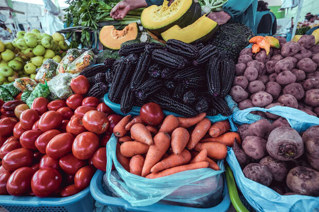 Fresh produce sits in baskets, displayed for sale. Vibrant red tomatoes, orange carrots, purple potatoes, and dark maize create a colorful arrangement. Halved squash and green apples add contrast.