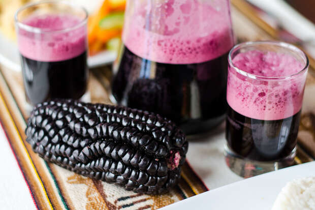 A glass pitcher and two glasses filled with purple juice and pink foam sit beside a black corn cob on a patterned tablecloth. Plates with food are partially visible.