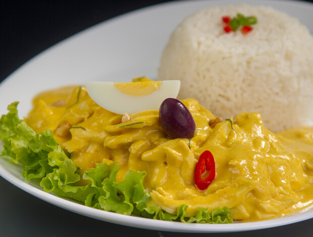 A dish of creamy yellow chicken stew garnished with lettuce, hard-boiled egg quarter, olive, and red chili. Served alongside a mound of white rice with red and green garnish.