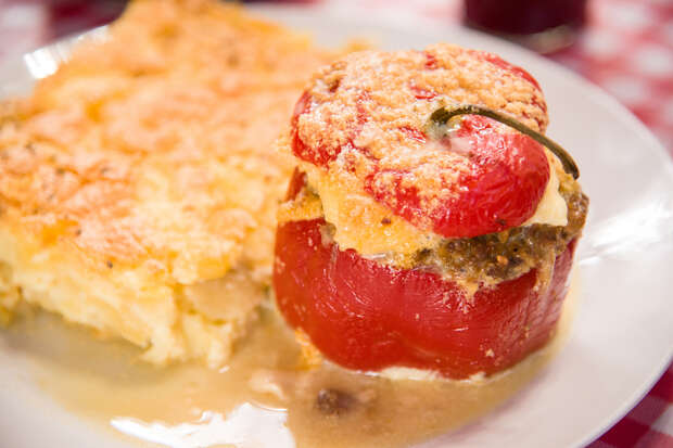 Stuffed red bell pepper sits on a white plate beside creamy potato gratin, with a checkered tablecloth in the background. The pepper is topped with a crispy, golden breadcrumb layer.