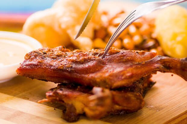 Fork and knife cutting into seasoned grilled meat on a wooden board, surrounded by crispy snacks and sauce, with blurred background.