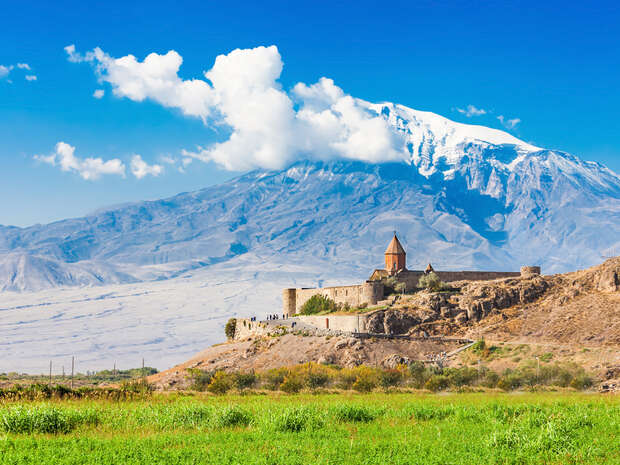 A historic monastery with a pointed roof sits atop a rocky hill, overlooking a green field. In the background, a snow-capped mountain rises under a vibrant blue sky.