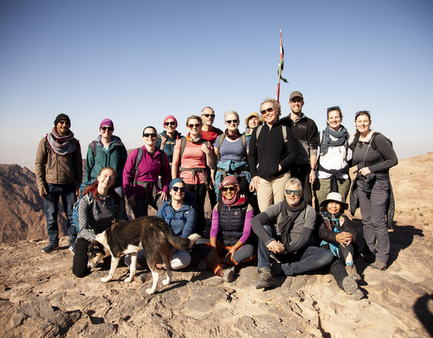 A diverse group of people stand together on a rocky mountaintop under a clear blue sky, some smiling at the camera, with a dog nearby and a flag waving in the background.