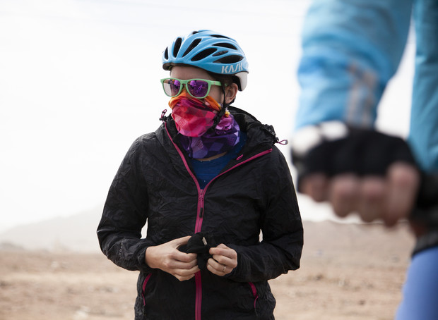 A person wearing a blue helmet and colorful scarf stands outdoors adjusting their jacket zipper, with another cyclist in a blue jacket nearby, in a dusty, open landscape.