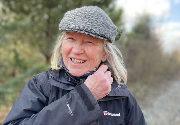 A person smiles while adjusting their collar with one hand. They wear a gray flat cap and black jacket labeled "Berghaus." Trees and a blue sky form the outdoor background.