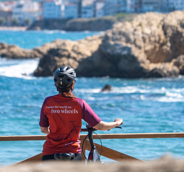 A person wearing a helmet and a red shirt with "Explore the world on two wheels" stands with a bicycle, facing a rocky coastline and a blue ocean.