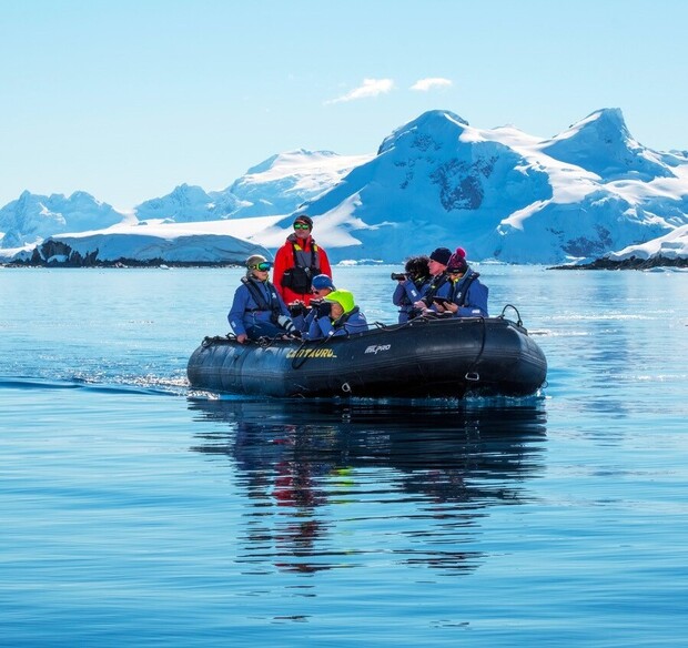 A black inflatable boat carries people in colorful gear, exploring calm waters surrounded by snow-covered mountains under clear blue skies.