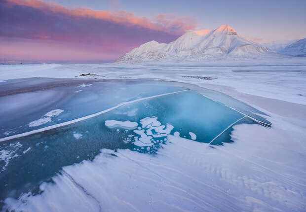 Frozen lake with icy patches set against a backdrop of snow-covered mountains under a pink-hued sky, creating a serene and cold arctic landscape.