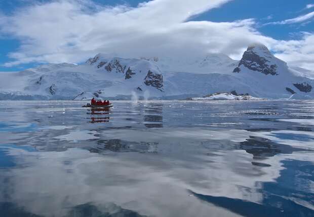 A small red boat carries people across calm, reflective waters, surrounded by snow-covered mountains under a partly cloudy blue sky.