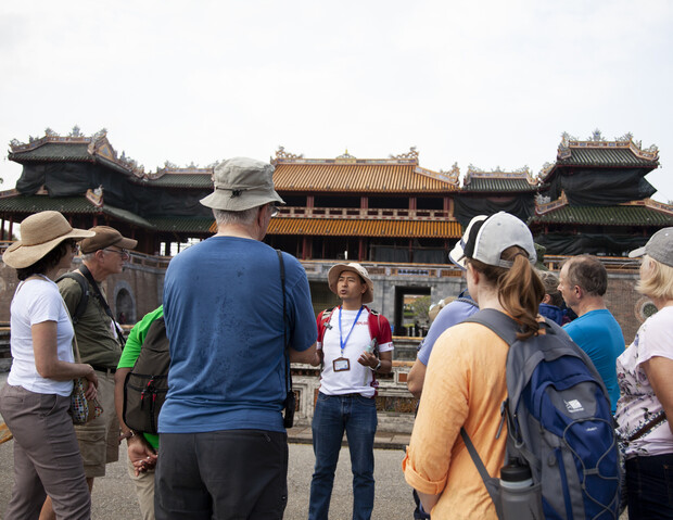 A tour guide, wearing a hat and badge, speaks to a group of tourists outside an ornate, traditional Asian palace with decorative roofs in a historical setting.