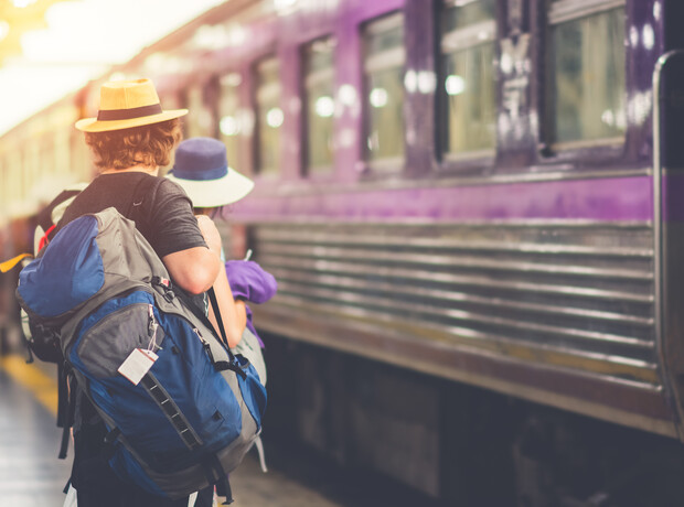 A person with a large backpack waits beside a purple train, wearing a straw hat and standing next to someone in a blue hat, at a train station platform.