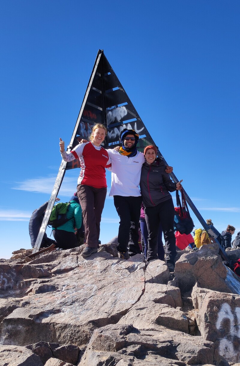 Three people stand smiling on a rocky mountaintop near a triangular metal sculpture covered in graffiti, under a clear blue sky with others in the background.