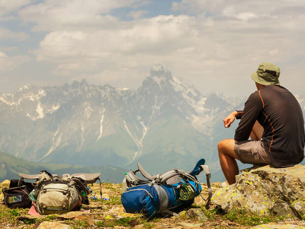 A person sits on a rocky outcrop, wearing a hat and outdoor clothing, gazing at distant snow-capped mountains. Several backpacks rest beside them, suggesting a hiking trip.