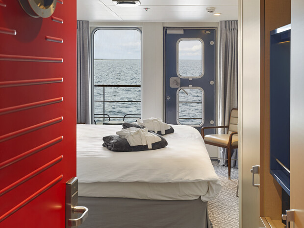 A neatly made bed with folded towels and robes is centered in a ship cabin, framed by a red door. Large windows reveal a calm ocean view, enhancing the cozy atmosphere.
