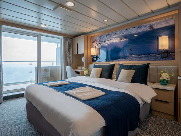 Luxurious bedroom features a neatly made bed with blue accents, positioned under an ocean-themed artwork. Large windows reveal a balcony with a sea view, creating a serene, modern ambience.