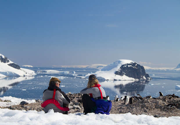 Two people in winter gear sit on a snowy shore observing penguins near an icy ocean, surrounded by snow-covered mountains under a clear blue sky.