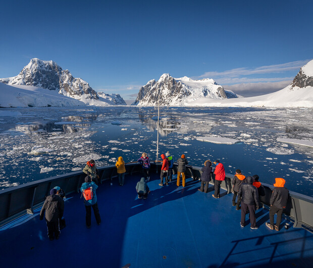 People in colorful jackets stand on a ship's blue deck, observing a serene, icy landscape with snow-covered mountains and scattered ice floes under a clear blue sky.