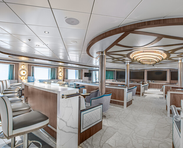 Luxurious interior features a sleek bar counter with high-backed stools, surrounded by plush seating and tables. The space is adorned with decorative lighting and wooden accents, creating an upscale lounge atmosphere.