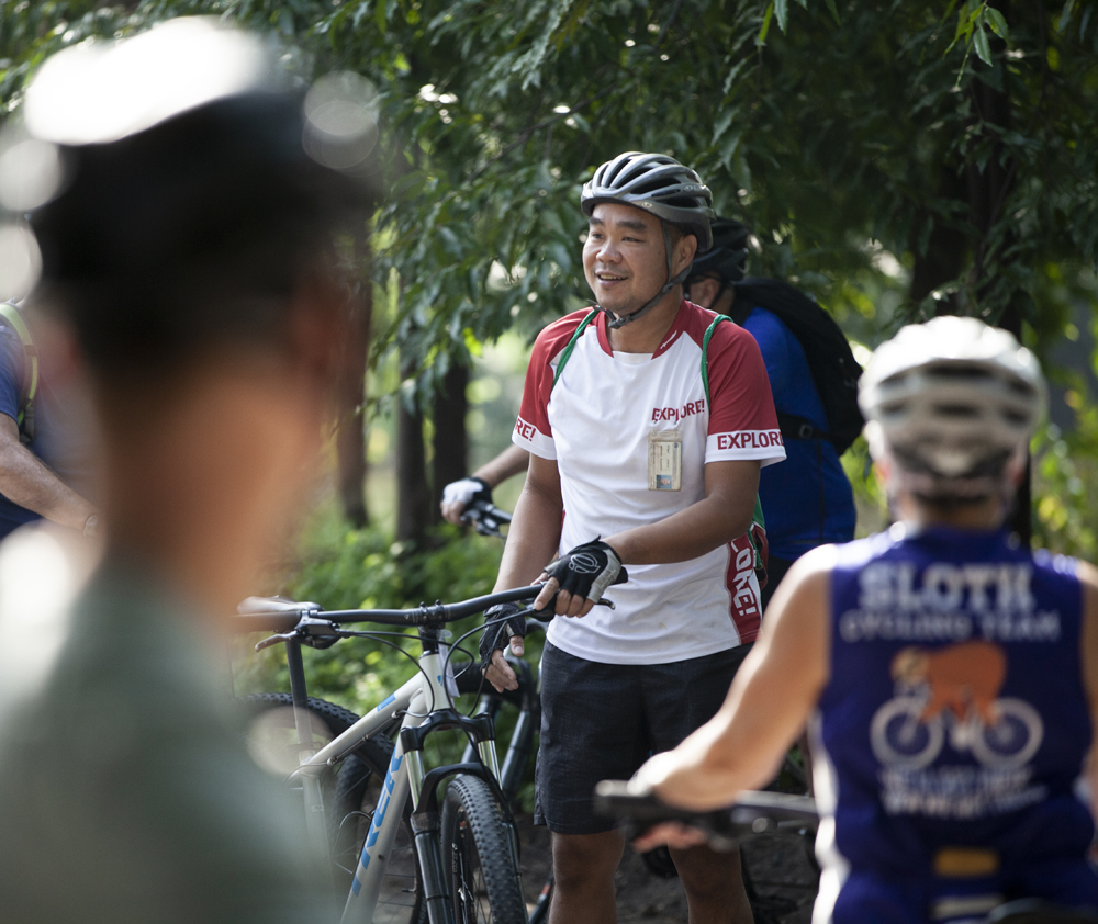 A cyclist wearing a helmet and red and white jersey labeled "EXPLORER" stands holding a bike, engaging with others in an outdoor park setting among trees.