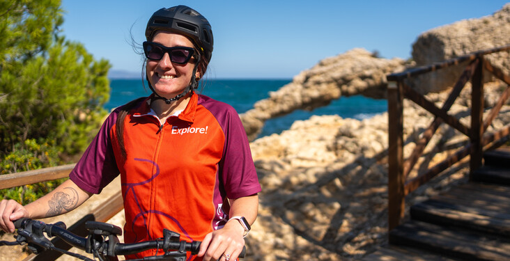 A person wearing a helmet and "Explore!" jersey smiles while holding a bicycle. They stand on a wooden walkway beside rocky terrain, with the ocean visible in the background.