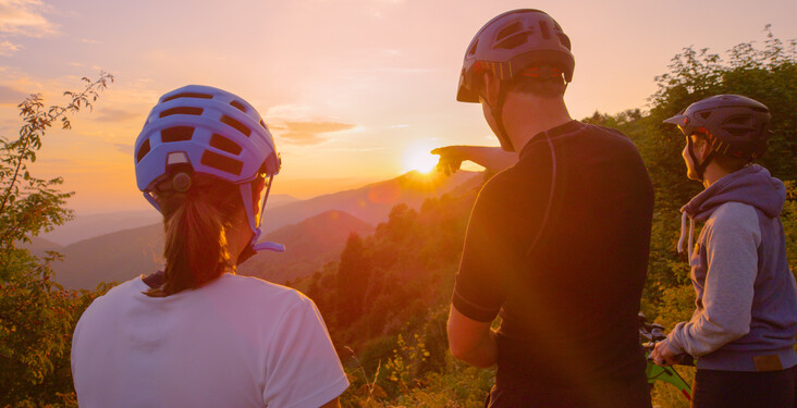 Three cyclists wearing helmets and casual clothing stand on a mountain trail. One points towards the scenic sunset over forested hills, creating a warm, picturesque outdoor setting.