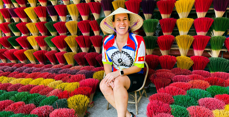 A person wearing a conical hat and colorful shirt, sitting joyfully among arranged bundles of vibrant incense sticks of red, yellow, and green, displayed against a dense backdrop.