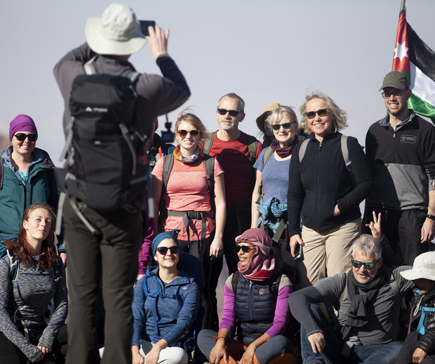 A group of hikers poses for a photo, taken by a person wearing a hat and backpack, against a clear sky. A flag is visible in the background.