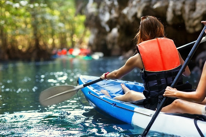 Two people paddle a blue kayak through clear water in a forested area, wearing life vests. In the background, blurred kayakers are visible near rocky outcrops.