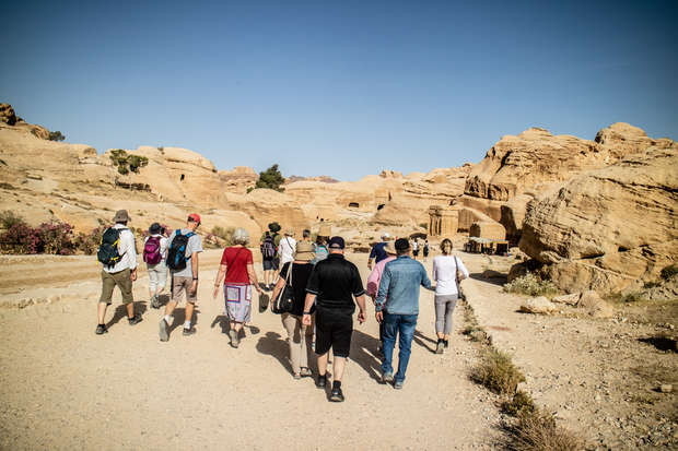 A group of people walks along a sandy path, surrounded by ancient, rocky desert formations under a clear blue sky.