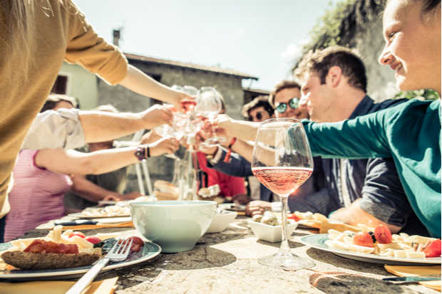 A group of people shares a toast with wine glasses around a sunlit outdoor table laden with plates of food in a relaxed, social setting.