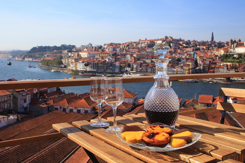 Decanter with red wine and two empty glasses rest on a wooden table with pastries, overlooking a scenic river and red-roofed town on a sunny day.