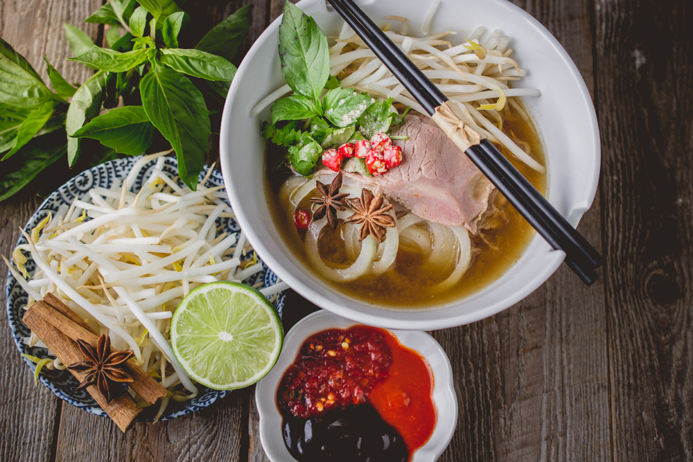A bowl of pho with beef, bean sprouts, herbs, chili, and star anise, served with chopsticks. Accompanied by fresh lime, herbs, and sauces on a wooden table.