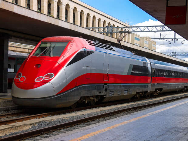 A sleek, modern high-speed train with a red and silver design is stationary on railway tracks at a train station, with a concrete platform and arched building in the background.