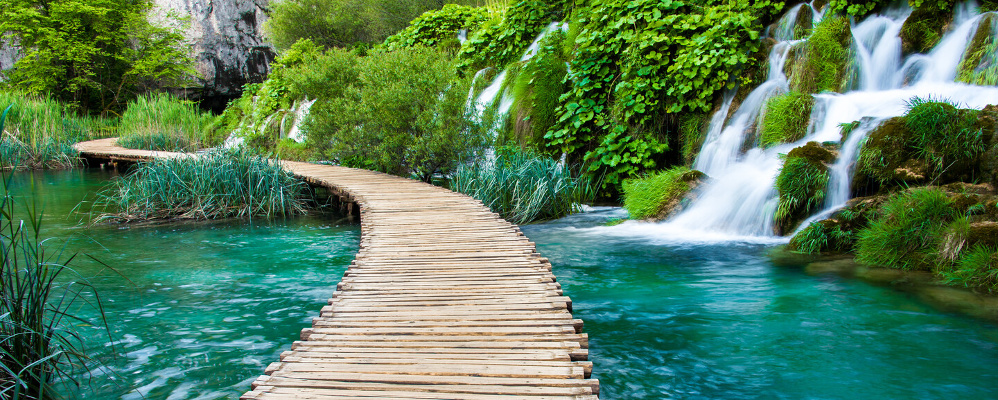 A wooden boardwalk curves over clear turquoise water, surrounded by lush greenery and cascading waterfalls nestled in a scenic, verdant landscape.