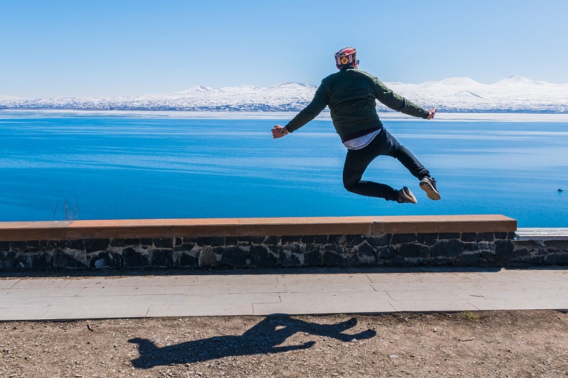 A person leaps joyfully in mid-air over a stone path, with a vast blue lake and snow-capped mountains under a clear sky in the background.