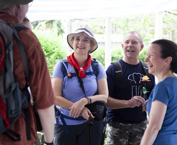 Several people, wearing casual outdoor clothing and backpacks, stand together smiling and talking in a garden setting with greenery around them. One person holds a flower.