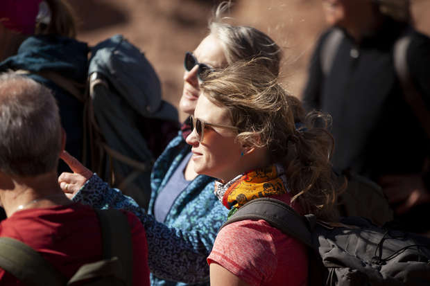 A group of hikers, wearing backpacks and sunglasses, stand outdoors, possibly on a trail. One person points ahead, perhaps indicating a path or landmark, under a sunny, scenic environment.