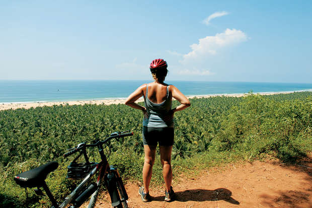 A person stands with hands on hips, wearing a red helmet, overlooking a coastal landscape of palm trees and ocean, with a bicycle leaning nearby on a dirt path.