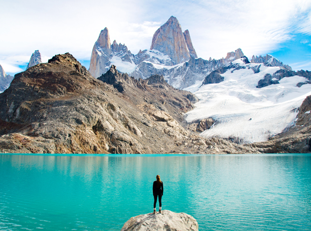 A person stands on a rock, facing a turquoise lake with a backdrop of rugged mountains and snow, under a partly cloudy sky.