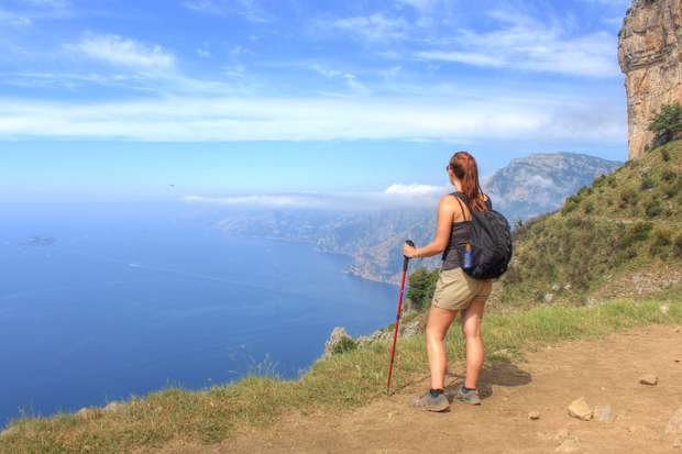 A person stands with a hiking pole, overlooking a vast ocean from a grassy cliffside trail, with mountains and a partly cloudy sky in the distance.